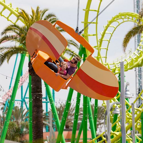 Two people enjoying a colorful amusement ride with green roller coaster tracks and palm trees in the background.